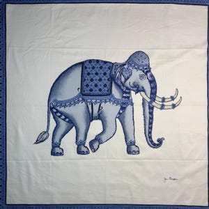 May include: A white textile with a blue elephant design. The elephant is decorated with blue patterns and a saddle. The textile's border is blue with a repeating pattern. The artist's signature is in the bottom right corner.