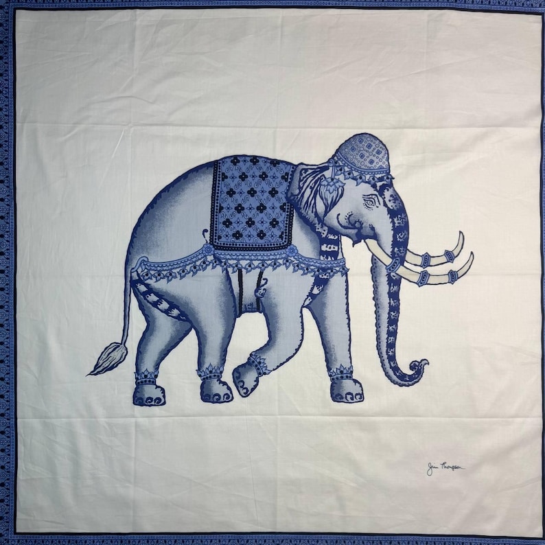 May include: A white textile with a blue elephant design. The elephant is decorated with blue patterns and a saddle. The textile's border is blue with a repeating pattern. The artist's signature is in the bottom right corner.