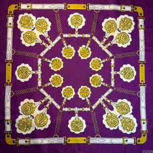 May include: A square purple scarf with a repeating pattern of gold and white decorative elements. The design includes stylized floral shapes, chains, and belt-like structures, creating a symmetrical and ornate aesthetic.