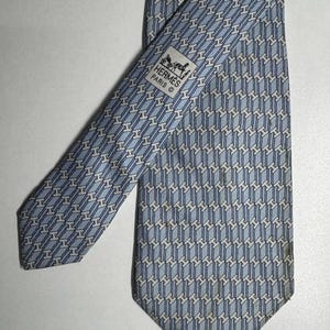 Pode incluir: Uma gravata de seda azul clara com um padrão geométrico repetido em branco. A gravata apresenta o logotipo "Hermès Paris" em uma pequena etiqueta. A gravata está em ângulo sobre uma superfície branca.