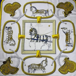 Puede incluir: Pañuelo de seda blanco con borde azul y amarillo, con una ilustración central de un carruaje tirado por caballos e ilustraciones de caballos enmarcadas. El pañuelo incluye el texto "GRAND APPARAT" y "HERMES-PARIS".