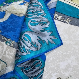 May include: Silk scarf in shades of blue, teal, and cream. The design incorporates classical figures and silver leaf patterns. The edges are a deep blue, with an intricate and detailed overall pattern.