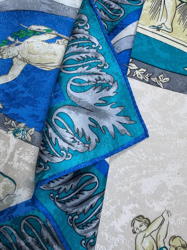 May include: Silk scarf in shades of blue, teal, and cream. The design incorporates classical figures and silver leaf patterns. The edges are a deep blue, with an intricate and detailed overall pattern.