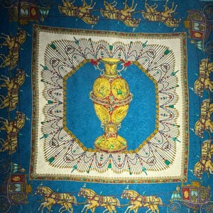 May include: Square silk scarf with a central design of a gold urn on a blue background, framed by a white border with ornate details. The scarf features horse-drawn carriages and decorative elements in gold, red, and green.