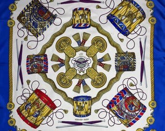 May include: A square silk scarf with a white central design of drums, drumsticks, and rope. The scarf has a blue border and gold accents. The text "LES TAMBOURS" is visible in the centre. The brand name "HERMÈS - PARIS" is also visible.