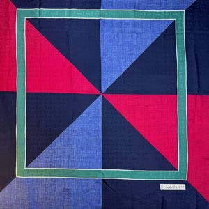 May include: A square silk scarf with a geometric design. The scarf features a pattern of triangles in shades of blue, red, and navy, framed by a green border with a white accent. The Yves Saint Laurent logo is visible in the corner.