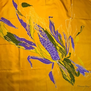 May include: A square, golden-yellow silk scarf with a botanical design. The design features a stylized corn plant with purple, green, and white accents. The scarf has the Christian Dior signature in the bottom right corner.