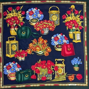 May include: A navy blue silk scarf featuring a repeating pattern of floral arrangements, handbags, and ornate clocks. The flowers are red, yellow, and blue. The handbags are green and red. The clocks are gold. The scarf has a red and gold border. The word "LANCEL" is printed on the bottom right.
