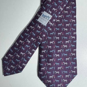 Pode incluir: Gravata de seda roxa com um padrão repetido de cavalos e cavaleiros brancos e azuis. A gravata tem uma etiqueta que diz "HERMES PARIS". Um acessório clássico.