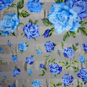 May include: A fabric with a floral pattern featuring large, vibrant blue roses and green leaves against a light gray background. The roses vary in size and are scattered across the fabric, creating a visually appealing design.