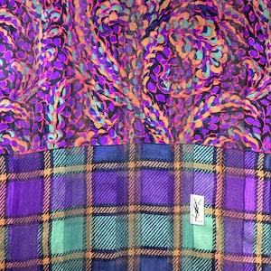 May include: A vibrant silk scarf featuring two distinct patterns. The top portion showcases a swirling design in shades of purple, pink, and teal. The bottom half displays a plaid pattern in purple, teal, and gold. A small logo is visible.