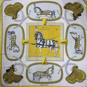 Puede incluir: Pañuelo cuadrado de seda con fondo blanco y panel central amarillo con un carruaje tirado por caballos. El diseño incluye ilustraciones enmarcadas de caballos y sillas de montar, con el texto "GRAND APPARAT" y "HERMES-PARIS".