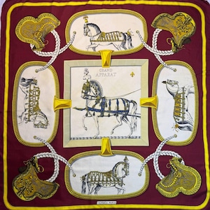 Puede incluir: Un pañuelo cuadrado de seda burdeos con borde dorado. El pañuelo presenta ilustraciones enmarcadas de caballos con arneses y monturas ornamentadas, y el texto "GRAND APPARAT". Un accesorio de lujo.