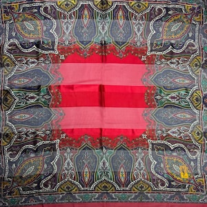 May include: A square, patterned scarf with a central red and pink striped design. The border features a paisley pattern in shades of blue, green, yellow, and purple. A small yellow logo is visible in the corner. The scarf is likely made of silk or a similar lightweight fabric.