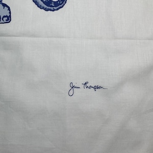 May include: White fabric featuring a blue and white ornate design. The fabric has a blue border with a repeating pattern. The signature "Jim Thompson" is written in blue script on the fabric.