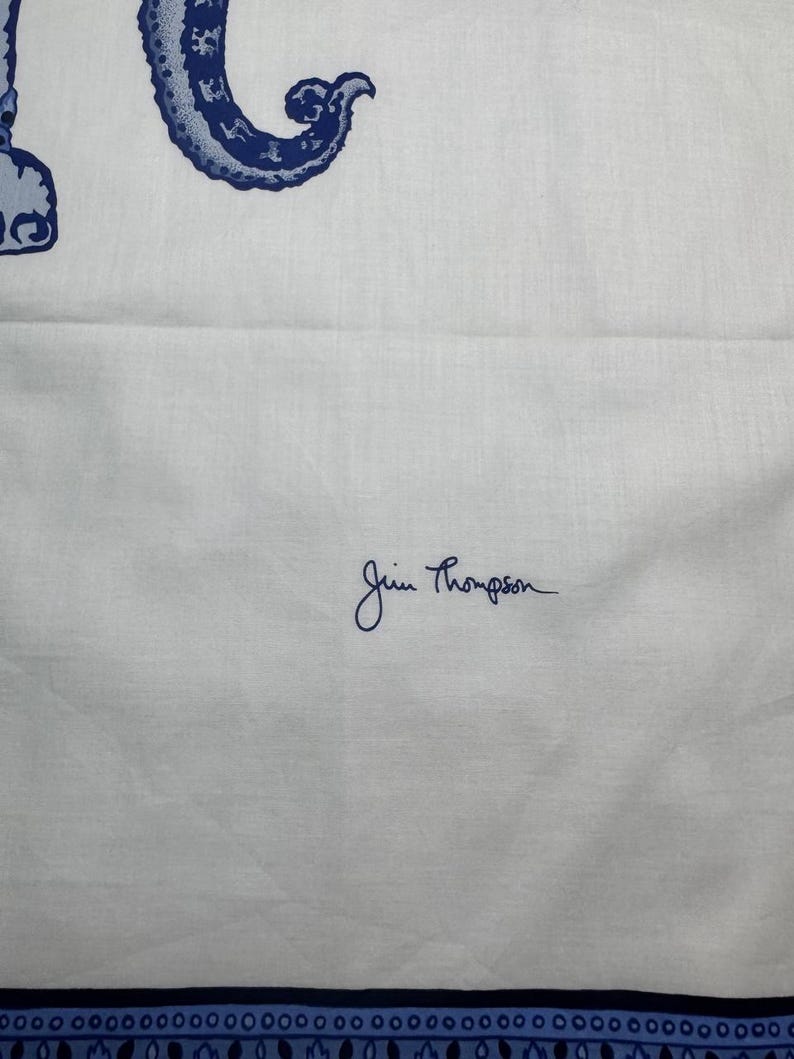 May include: White fabric featuring a blue and white ornate design. The fabric has a blue border with a repeating pattern. The signature "Jim Thompson" is written in blue script on the fabric.