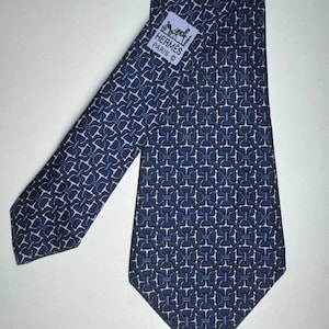 Pode incluir: Uma gravata azul marinho com um padrão geométrico repetitivo em branco e azul claro. A gravata apresenta uma etiqueta com o texto "HERMES PARIS" e um logótipo. A gravata é feita de um tecido liso.
