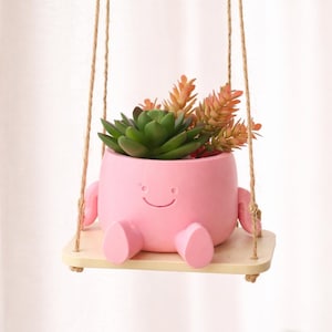Puede incluir: Una maceta rosa con una cara sonriente, brazos y piernas, suspendida por una cuerda. La maceta contiene suculentas verdes y naranjas y se asienta sobre un pequeño columpio de madera. Un artículo decorativo encantador y caprichoso.