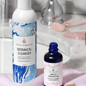 May include: Two skincare products: a white and blue botanical cleanser bottle and a small blue makeup removing oil bottle. The cleanser bottle has a white cap and a label with the text "BOTANICAL CLEANSER". The oil bottle has a dropper. Both bottles are on a pink towel.
