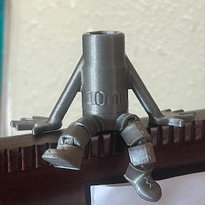 May include: A gray 3D printed figurine of a person sitting on a ledge. The figurine has a 10mm socket on its torso and articulated arms and legs.