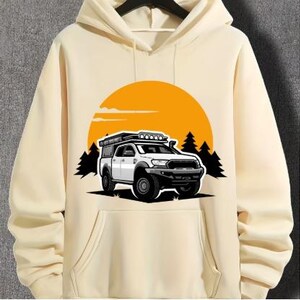 Puede incluir: Una sudadera con capucha de color crema con un gráfico de una camioneta blanca con una baca en el techo conduciendo por un paisaje de bosque con un sol amarillo en el fondo.
