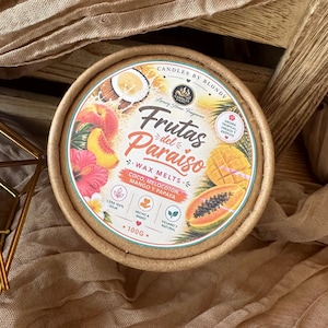 Op de afbeelding: Een ronde kartonnen verpakking met wax melts, gelabeld als Frutas del Paraiso. Het etiket bevat illustraties van kokosnoot, perzik, mango, papaja en hibiscusbloemen. De tekst bevat Candles by Blondy, 100g en iconen voor sojawas en handgemaakt.