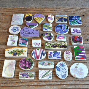 May include: A collection of assorted ceramic shards and tiles in various shapes and sizes, featuring floral patterns, fruit motifs, and a purple piece with the word Peace written in black script, including an Imperial Plums label.