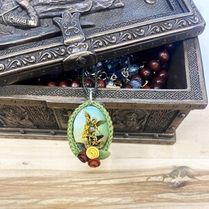 May include: A green crocheted oval pendant with a picture of an angel inside. The pendant has a small cluster of yellow and red flowers attached to the bottom.