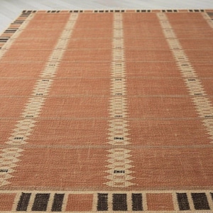 May include: A rectangular rug in terracotta with vertical cream-coloured geometric stripes. The rug features a border of alternating brown and tan squares. The rug is made of a woven material, suitable for a living space.