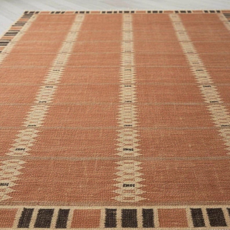 May include: A rectangular rug in terracotta with vertical cream-coloured geometric stripes. The rug features a border of alternating brown and tan squares. The rug is made of a woven material, suitable for a living space.