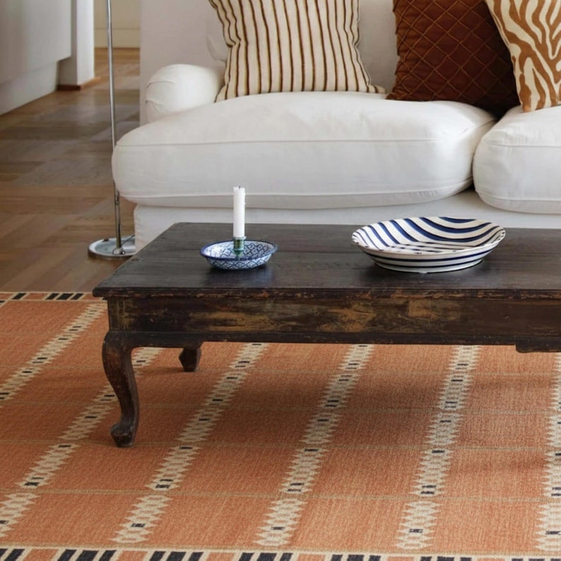 May include: A living room scene with a dark wooden coffee table holding a candle and a bowl with blue stripes. An orange and white patterned rug is in the foreground. A white sofa with striped and patterned cushions is visible.