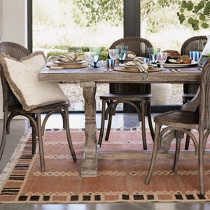 May include: A dining table set for a meal, with wicker chairs and a patterned rug. The wooden table is laden with plates, glasses, and napkins. A decorative pillow sits on a chair. The scene suggests a relaxed, inviting dining experience, with natural light.