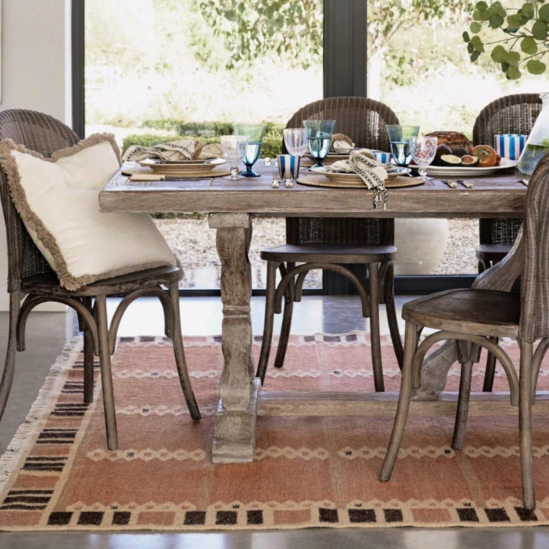 May include: A dining table set for a meal, with wicker chairs and a patterned rug. The wooden table is laden with plates, glasses, and napkins. A decorative pillow sits on a chair. The scene suggests a relaxed, inviting dining experience, with natural light.