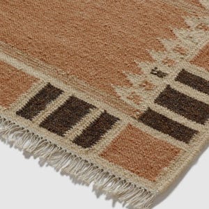 May include: Close-up of a woven rug with a geometric pattern. The rug has a beige background with brown and tan geometric shapes. The edges are fringed. The rug is made of a textured material.
