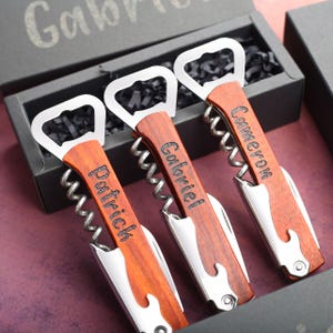 Puede incluir: Tres abridores de botellas personalizados con mangos de madera y nombres grabados: Patrick, Gabriel y Cameron. Los abridores de botellas están en una caja negra con papel triturado negro.