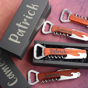 Pode incluir: Três abridores de garrafas personalizados com saca-rolhas em caixas pretas. Os abridores de garrafas têm alças de madeira com os nomes "Patrick", "Cameron" e "Gabriel" gravados neles.