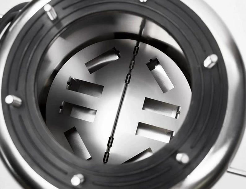 May include: A circular stainless steel chamber featuring a central hinged plate with rectangular cutouts. The interior is surrounded by a thick black rubber gasket secured with metal fasteners, creating a mechanical industrial appearance.