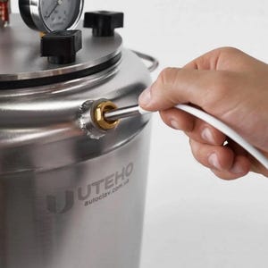 May include: A stainless steel autoclave with a pressure gauge and black knobs on the lid. The side of the cylindrical vessel features a brass fitting with a white cable inserted. The brand name UTEHO and the website autoclav.com.ua are printed.