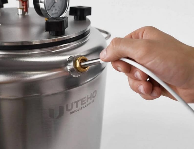 May include: A stainless steel autoclave with a pressure gauge and black knobs on the lid. The side of the cylindrical vessel features a brass fitting with a white cable inserted. The brand name UTEHO and the website autoclav.com.ua are printed.