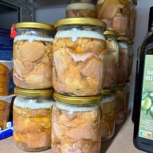 May include: Several clear glass jars with gold-coloured metal lids, each filled with chunks of preserved meat submerged in a layer of rendered fat. The jars are stacked in a group, showing the golden-yellow fat and pieces of cooked meat inside.