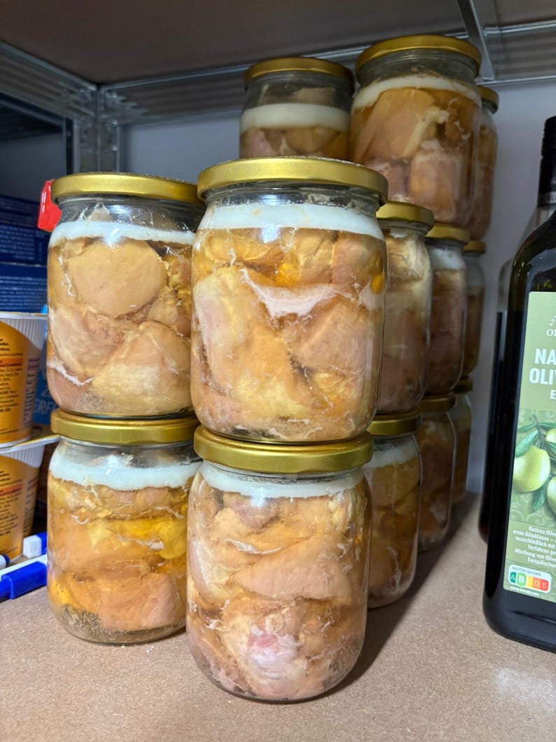 May include: Several clear glass jars with gold-coloured metal lids, each filled with chunks of preserved meat submerged in a layer of rendered fat. The jars are stacked in a group, showing the golden-yellow fat and pieces of cooked meat inside.