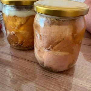 May include: Two clear glass jars with gold-coloured metal lids, each filled with preserved food items submerged in a translucent liquid. The contents appear to be solid, light-coloured pieces of meat or fat, showing distinct textures through the glass.