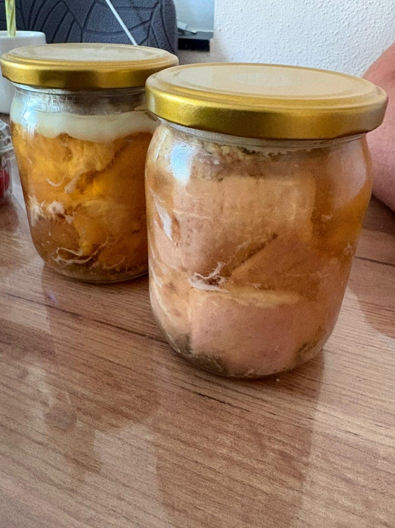 May include: Two clear glass jars with gold-coloured metal lids, each filled with preserved food items submerged in a translucent liquid. The contents appear to be solid, light-coloured pieces of meat or fat, showing distinct textures through the glass.