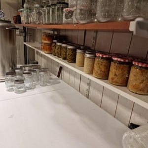 May include: A large stainless steel pressure canner sits on a white surface next to several empty glass jars with metal lids. Above, white shelves hold rows of filled glass jars containing preserved vegetables and food with various metal lids.