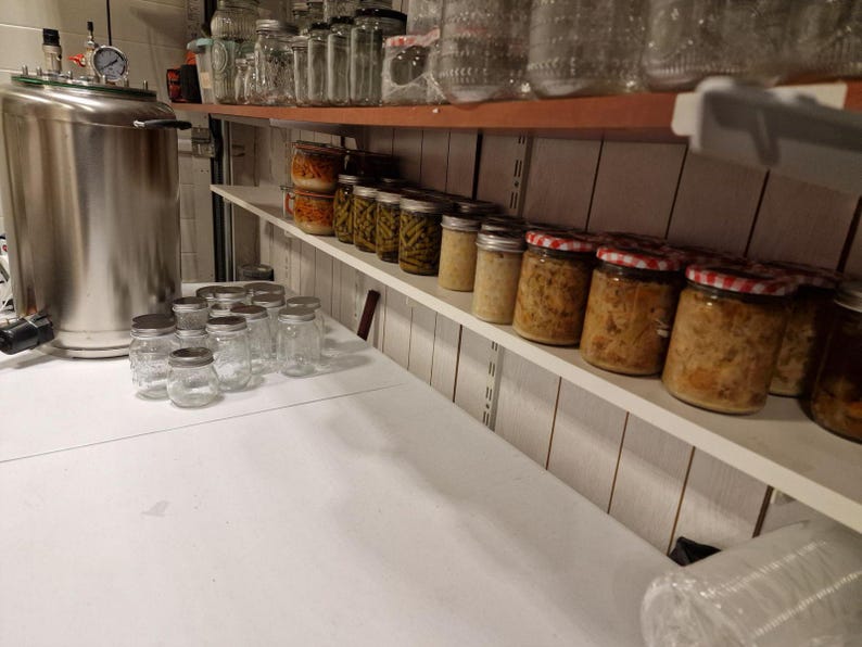 May include: A large stainless steel pressure canner sits on a white surface next to several empty glass jars with metal lids. Above, white shelves hold rows of filled glass jars containing preserved vegetables and food with various metal lids.