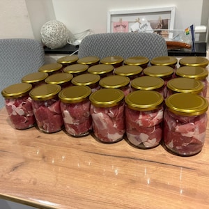 May include: A collection of twenty-four clear glass jars filled with raw, cubed red meat and white fat. Each jar is sealed with a circular, metallic gold-coloured lid. The jars are arranged in a grid formation on a light brown wooden surface.