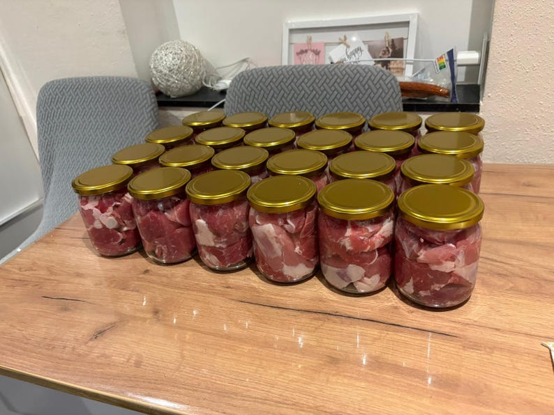 May include: A collection of twenty-four clear glass jars filled with raw, cubed red meat and white fat. Each jar is sealed with a circular, metallic gold-coloured lid. The jars are arranged in a grid formation on a light brown wooden surface.