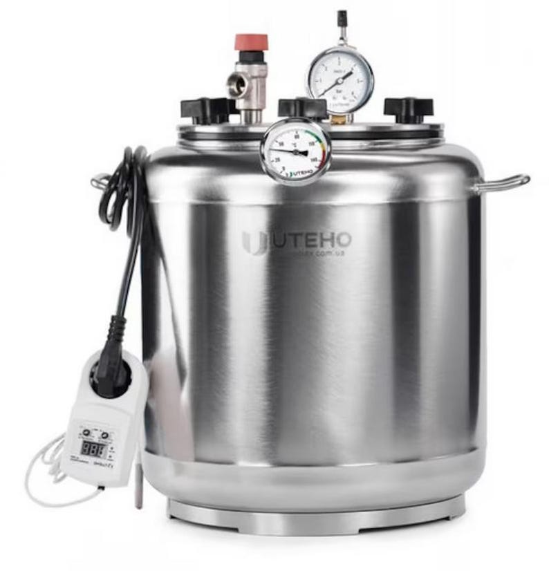 May include: A stainless steel autoclave with a cylindrical body, featuring a top lid secured by black knobs. It includes a pressure gauge, a temperature gauge, a red valve, and a digital controller with a power cord attached to the side.