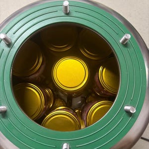 May include: A stainless steel canning or processing device with a circular green gasket featuring four protruding metal bolts. Inside the device, multiple glass jars with bright gold lids are arranged in a circular pattern. The word UTEHO is embossed on the green rim.