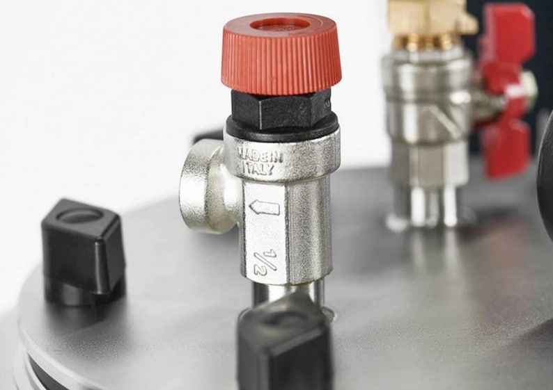 May include: A metallic pressure relief valve with a red plastic cap, featuring the text MADE IN ITALY and a 1/2 inch marking. The valve is mounted on a circular stainless steel plate alongside a brass fitting with a red handle and black knobs.